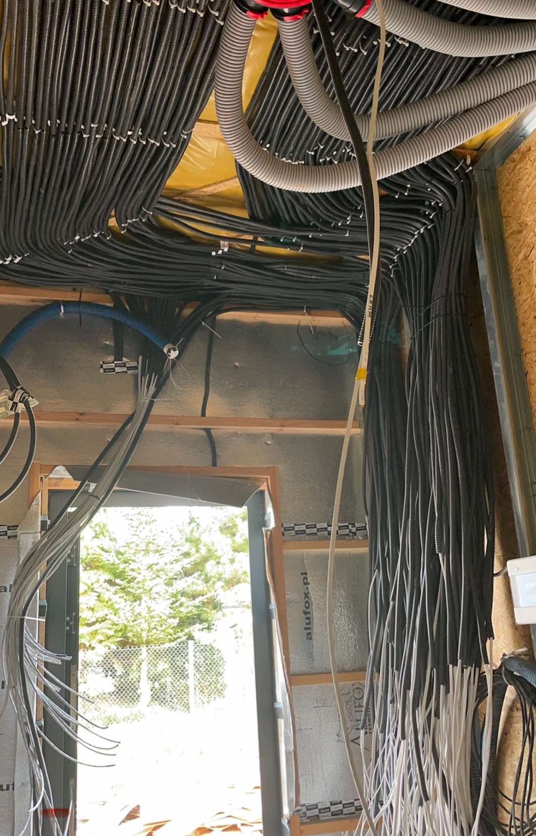 Sufit i fragment ściany w trakcie instalacji elektrycznej: liczne czarne i szare rury karbowane prowadzone równolegle, kable wychodzące z rur, widoczne elementy konstrukcji budynku.