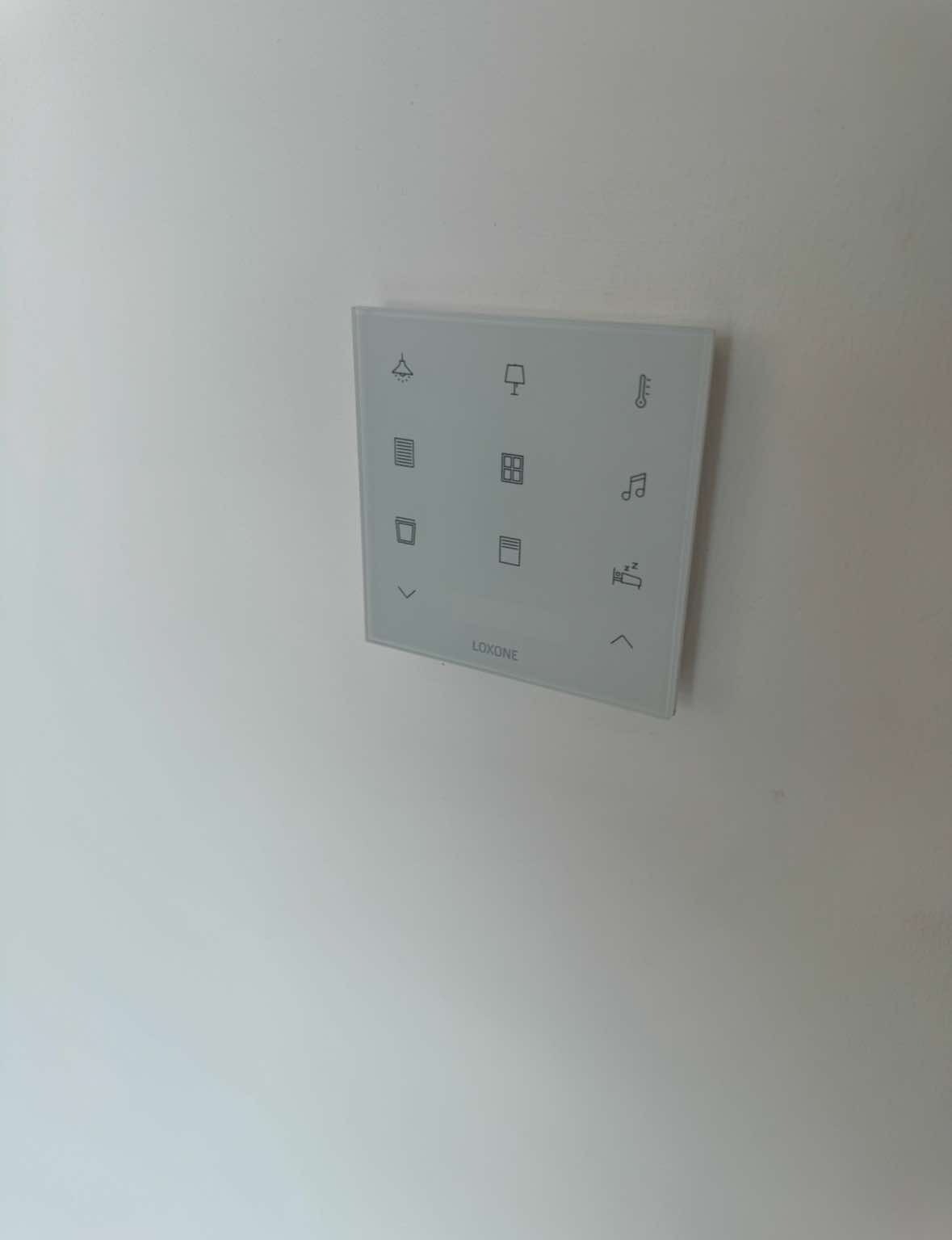 Biały panel sterowania systemu Loxone zamontowany na ścianie, z ikonami oświetlenia, rolet, temperatury i muzyki.