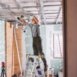 Pracownik w okularach ochronnych montuje metalowy stelaż sufitu podwieszanego, stojąc na składanej drabinie w pomieszczeniu z częściowo wykończonymi ścianami, widoczne elementy instalacji...