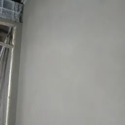 Świeżo nałożony, jasnoszary tynk na ścianie, widoczny fragment okna z surowym wykończeniem i niedoskonałościami przy suficie.