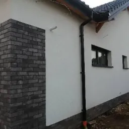 Fragment elewacji domu z ociepleniem i tynkiem strukturalnym, z czarną rynną spustową i fragmentem ściany wykończonej czarną cegłą klinkierową.
