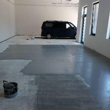 Szare, częściowo pomalowane betonowe podłoże w pustym pomieszczeniu z widocznym wiadrem i wałkiem malarskim w rogu, na tle zaparkowany niebieski samochód dostawczy i okna.