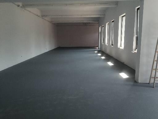 Przestronne, puste pomieszczenie z szarą podłogą, białymi ścianami i rzędem okien po prawej stronie, wpadające światło słoneczne, drabina oparta o ścianę w prawym dolnym rogu.