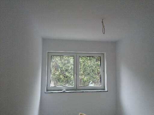 Wnętrze pokoju z białymi ścianami po malowaniu, widoczne okno z widokiem na zieleń, sufit z niedokończonym mocowaniem lampy.