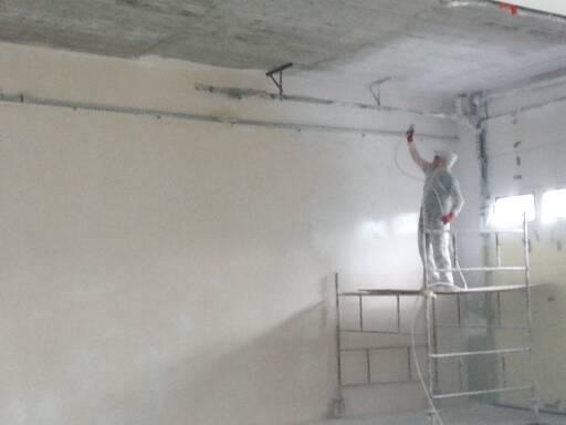 Pracownik w kombinezonie ochronnym maluje sufit i ścianę natryskowo z rusztowania, widoczne instalacje na suficie i ściany w stanie surowym.
