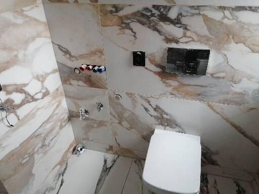 Nowoczesna toaleta podwieszana w łazience wyłożonej płytkami imitującymi marmur, widoczne instalacje wodno-kanalizacyjne i przyciski spłukujące.