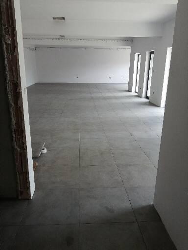 Przestronne wnętrze w trakcie prac wykończeniowych, z szarymi płytkami podłogowymi, białymi ścianami i widocznymi instalacjami na suficie, z fragmentem surowego muru przy wejściu.