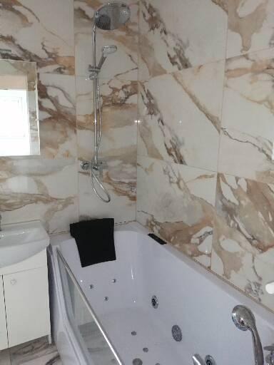 Biała wanna z hydromasażem w łazience wykończonej marmurowymi płytkami, widoczna bateria wannowa, kabina prysznicowa i umywalka z szafką.