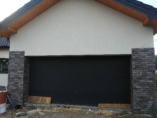 Elewacja garażu z czarną bramą segmentową, wykończona szarą cegłą klinkierową po bokach, z widocznym fragmentem ocieplonej ściany i drewnianym podbitką dachu.