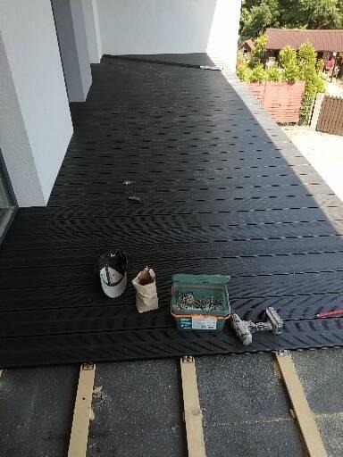 Instalacja czarnego tarasu kompozytowego na balkonie, widoczne legary, wkręty w zielonym pudełku, wkrętarka, czapka i worek cementu.