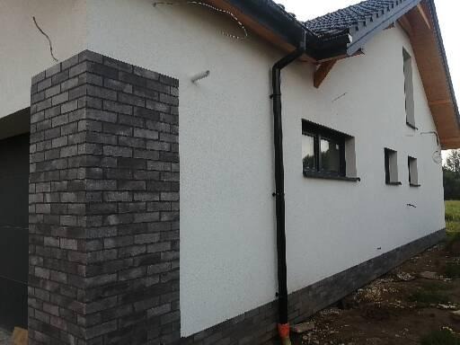 Fragment elewacji domu z ociepleniem i tynkiem strukturalnym, z czarną rynną spustową i fragmentem ściany wykończonej czarną cegłą klinkierową.