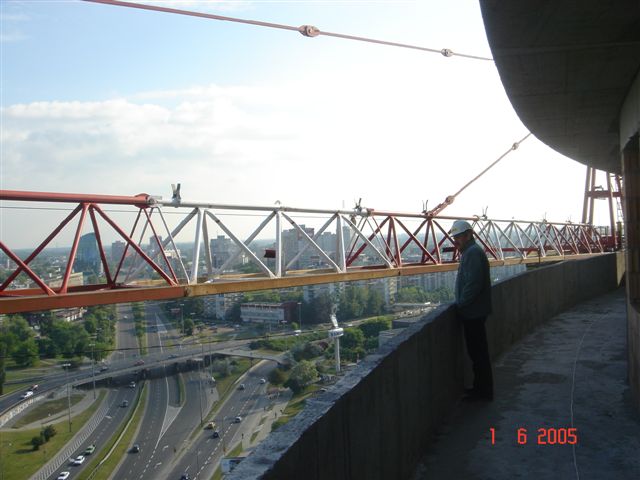 Pracownik w kasku stoi na betonowym balkonie wysokiego budynku w trakcie budowy, widoczna konstrukcja rusztowania i panorama miasta w tle.