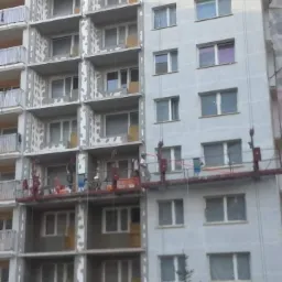 Wielopiętrowy budynek w trakcie prac elewacyjnych, widoczne rusztowania wiszące i pracownicy w kaskach, część ściany z oknami jest już pomalowana na szaro, a część przygotowana pod ocieplenie...