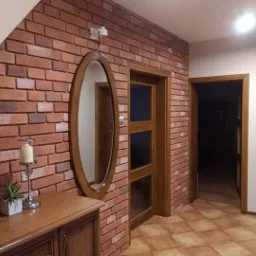 Korytarz z ceglaną ścianą, owalnym lustrem w drewnianej ramie, komodą z dekoracją oraz brązowymi płytkami podłogowymi i drewnianymi drzwiami.