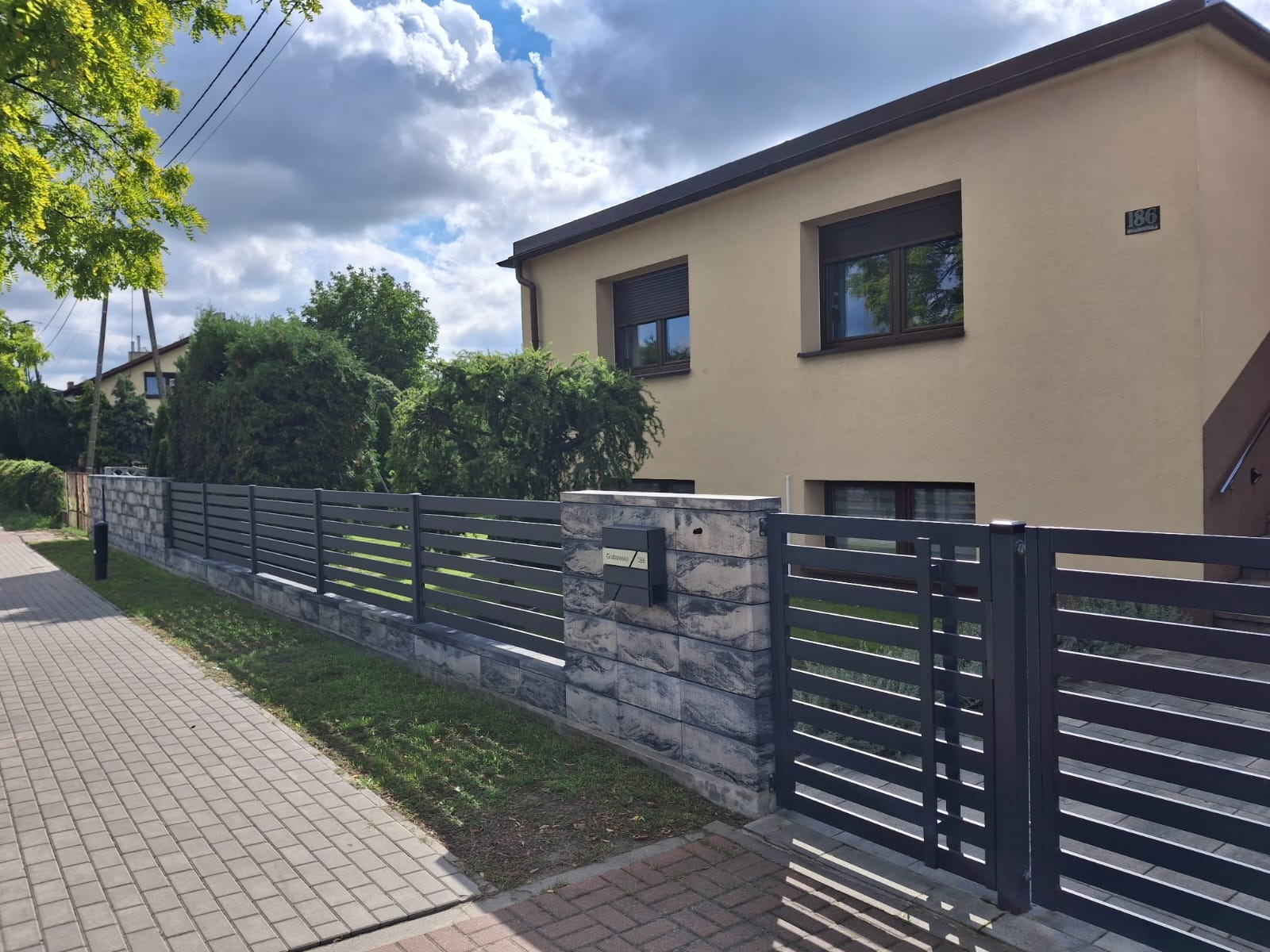 Nowoczesny dom z beżową elewacją i szarym, poziomym ogrodzeniem panelowym z kamiennymi słupkami, skrzynka pocztowa na jednym ze słupków, kostka brukowa.