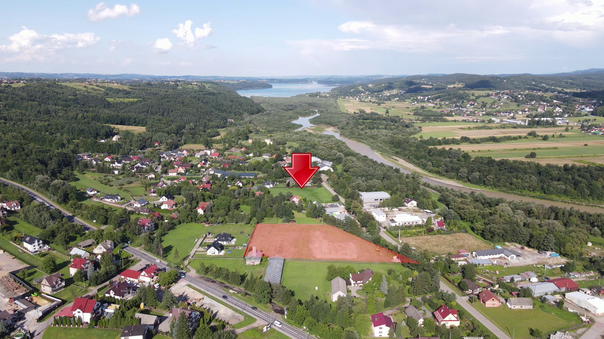 Szeroki plan z lotu ptaka na teren budowy osiedla w Myślenicach, z zaznaczonym obszarem przygotowanym pod inwestycję i czerwoną strzałką wskazującą na lokalizację. W tle widoczny krajobraz z rzeką...