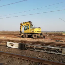 Żółta koparka Liebherr 900 na torach kolejowych, z ułożonymi podkładami kolejowymi w tle, widoczna infrastruktura kolejowa.