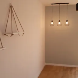 Jasne wnętrze z drewnianą podłogą, pomalowaną ścianą w odcieniach beżu i szarości, trzema lampami wiszącymi na czarnej listwie oraz dekoracyjnymi półkami wiszącymi na sznurkach.