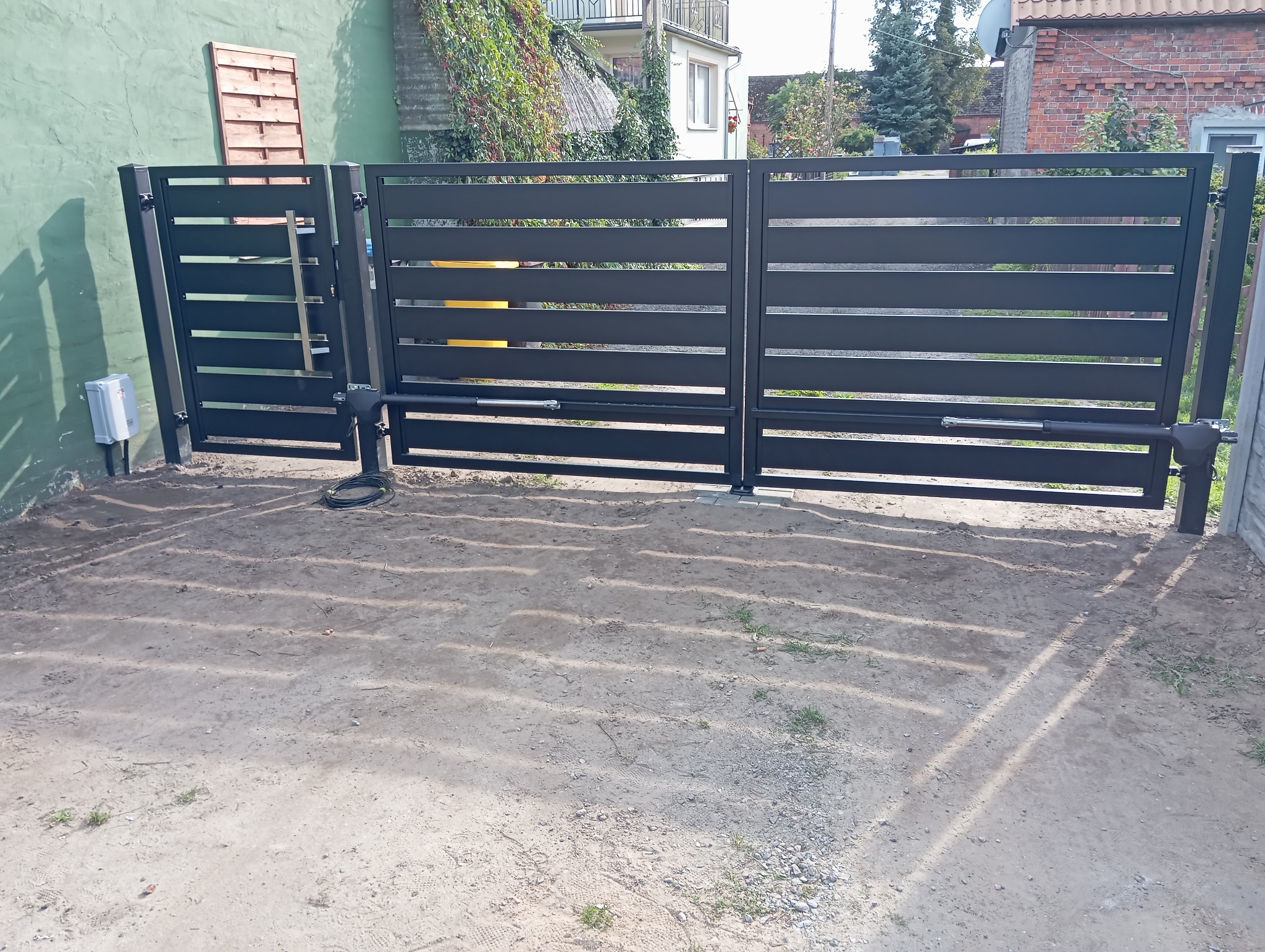 Nowoczesna, przesuwna brama z automatyką, z poziomymi panelami. Widoczne siłowniki i skrzynka sterownicza. Brama zamontowana na posesji z zielonym budynkiem w tle.