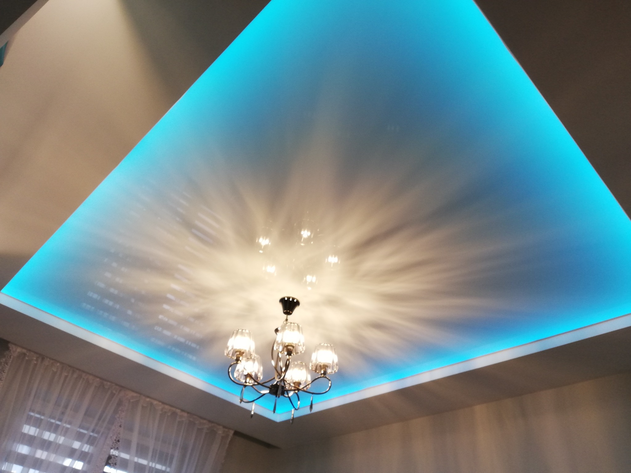 Podwieszany sufit z oświetleniem LED w kolorze turkusowym, z widocznym żyrandolem z kryształkami, odbijającym światło na suficie.