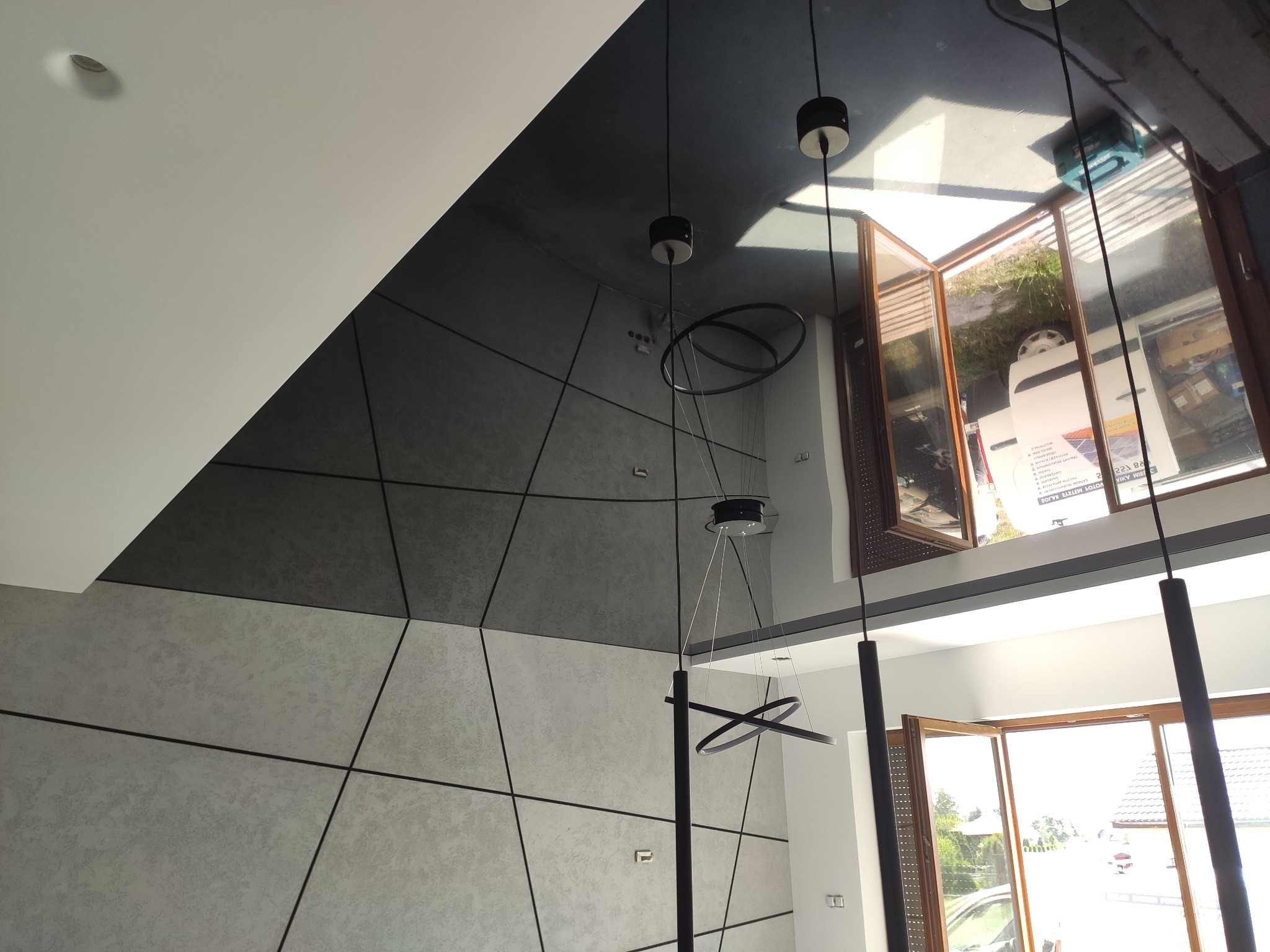 Wnętrze z nowoczesnym oświetleniem: lampy wiszące z okrągłymi elementami, widok z dołu na sufit z geometrycznym wzorem i duże okno z widokiem na zewnątrz.