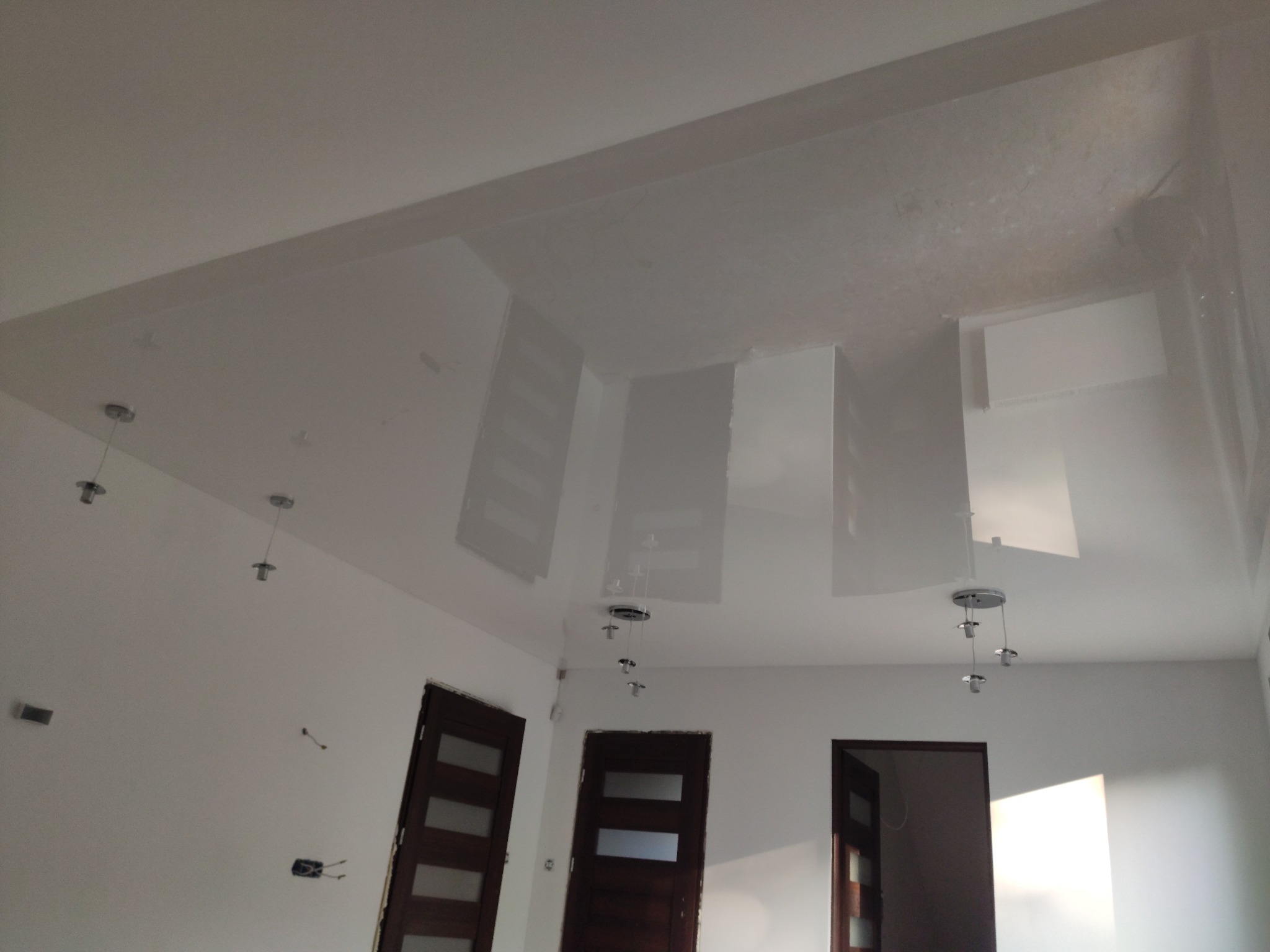 Biały, błyszczący sufit napinany z zamontowanymi lampami wiszącymi i widocznymi trzema parami drzwi z ciemnego drewna.