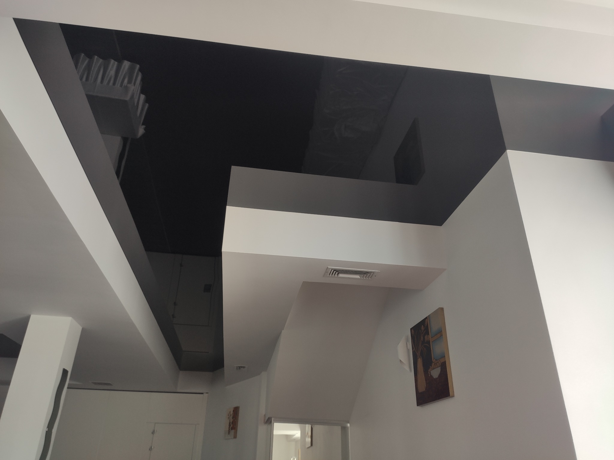 Nowoczesny sufit podwieszany z czarnym, błyszczącym panelem i wbudowaną kratką wentylacyjną, widok z dołu, z obrazem na ścianie w tle.