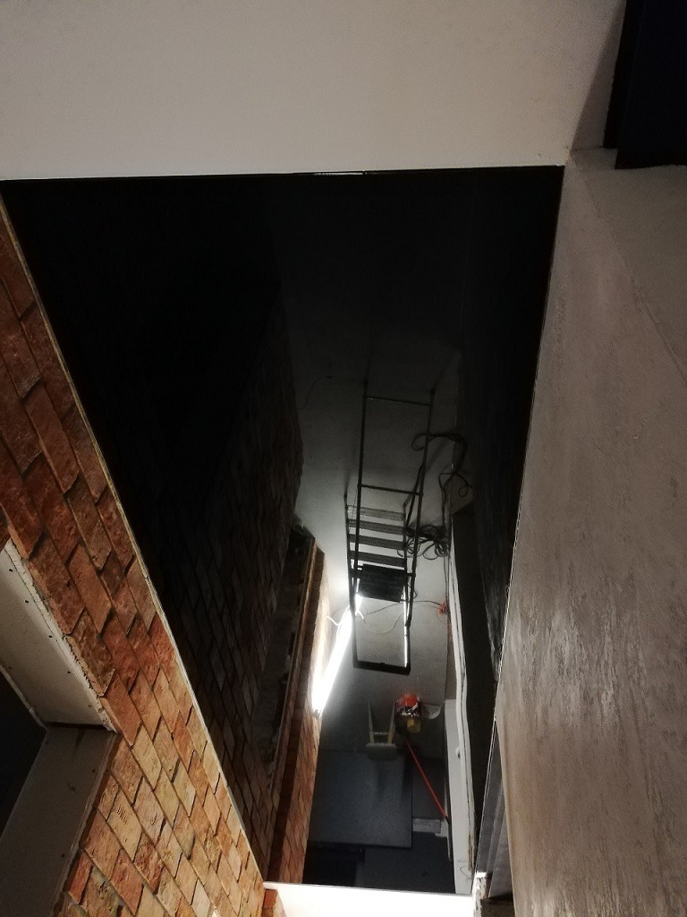 Wnętrze w trakcie montażu instalacji wentylacyjnej w Mielcu, widok z góry na drabinę i kanały wentylacyjne, surowa cegła na ścianie.