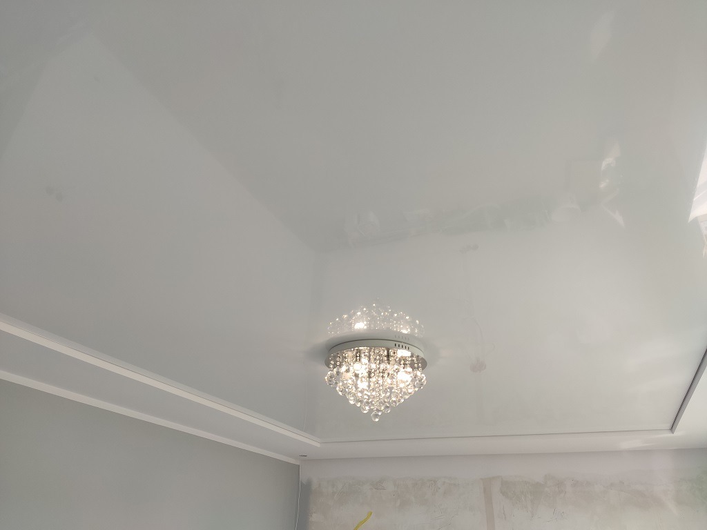 Biały, błyszczący sufit napinany z ozdobnym, kryształowym żyrandolem w centrum, widoczne listwy przypodłogowe i fragment ściany w trakcie wykończenia.
