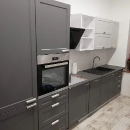 Szara zabudowa kuchenna z piekarnikiem i okapem, białe szafki wiszące, czarny zlew i bateria, podłoga imitująca drewno.