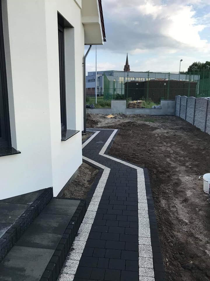 Ścieżka z ciemnej kostki brukowej z jasnymi obrzeżami prowadząca od schodów domu w Lesznie, w tle widoczny fragment ogrodzenia i budynek.
