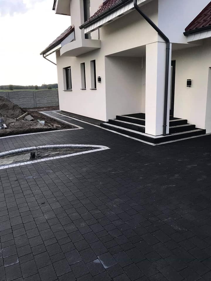 Ułożona czarna kostka brukowa przed nowoczesnym domem z białymi ścianami i czarnymi schodami wejściowymi, widoczny fragment niezakończonej obwódki z kostki granitowej.