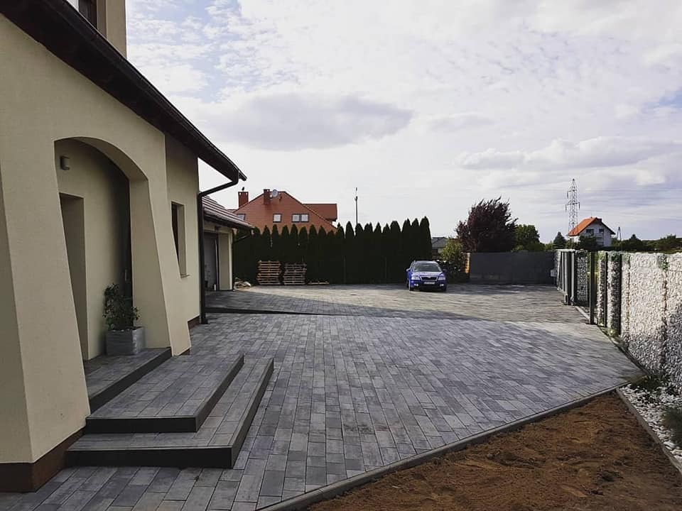 Nowoczesny podjazd z szarej kostki brukowej ułożonej w cegiełkę, z trzema stopniami wejściowymi do domu i widocznym samochodem na tle zielonych tuj.
