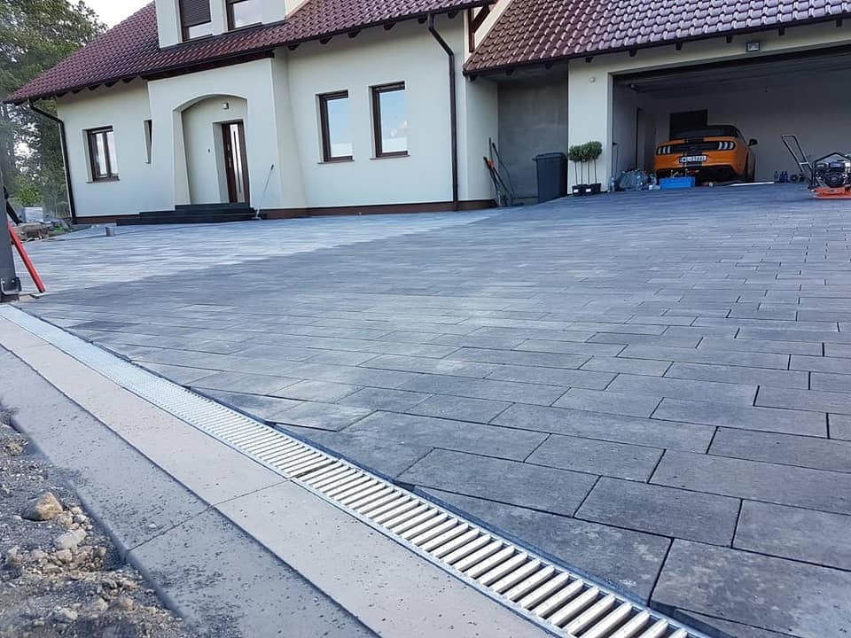Ułożona szara kostka brukowa na podjeździe z odpływem liniowym, w tle dom jednorodzinny z garażem i pomarańczowy samochód