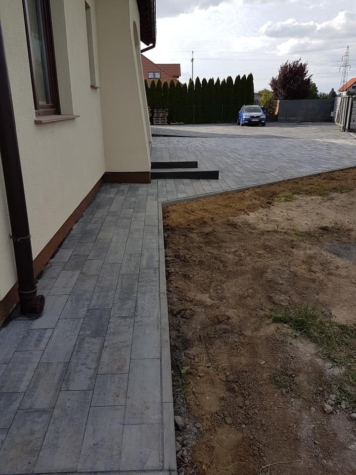 Ułożona szara kostka brukowa prowadząca od ściany domu do podjazdu, z widocznymi schodami wejściowymi i przygotowanym terenem pod trawnik.