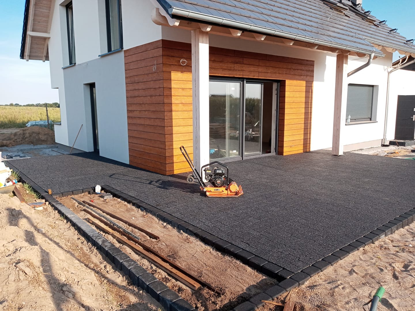 Częściowo ułożona czarna kostka brukowa na posesji przed domem z drewnianą elewacją. W tle widoczna zagęszczarka do kostki brukowej i przygotowane obrzeża.