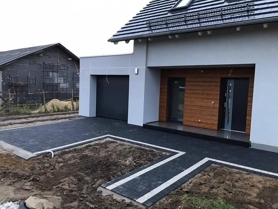 Nowoczesny dom jednorodzinny z ciemną kostką brukową na podjeździe, obrzeża z jasnej kostki, widoczne rabaty ziemne przed domem, budynek w trakcie wykończenia w tle.