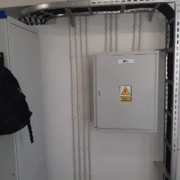 Szara skrzynka elektryczna RPW z ostrzeżeniem o napięciu, zamontowana na białej ścianie z pionowymi rurami w białych uchwytach, widoczna metalowa korytka kablowe pod sufitem i metalowa drabinka...