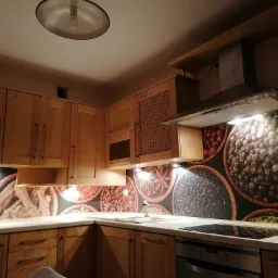 Kuchnia z jasnymi drewnianymi szafkami i tapetą przedstawiającą przyprawy w misach, oświetlona punktowymi lampkami podszafkowymi.