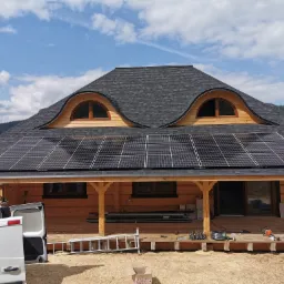 Drewniany dom z ciemną dachówką i zamontowanymi panelami fotowoltaicznymi; na werandzie widoczne narzędzia i materiały montażowe, w tle błękitne niebo z chmurami i zalesione wzgórza.