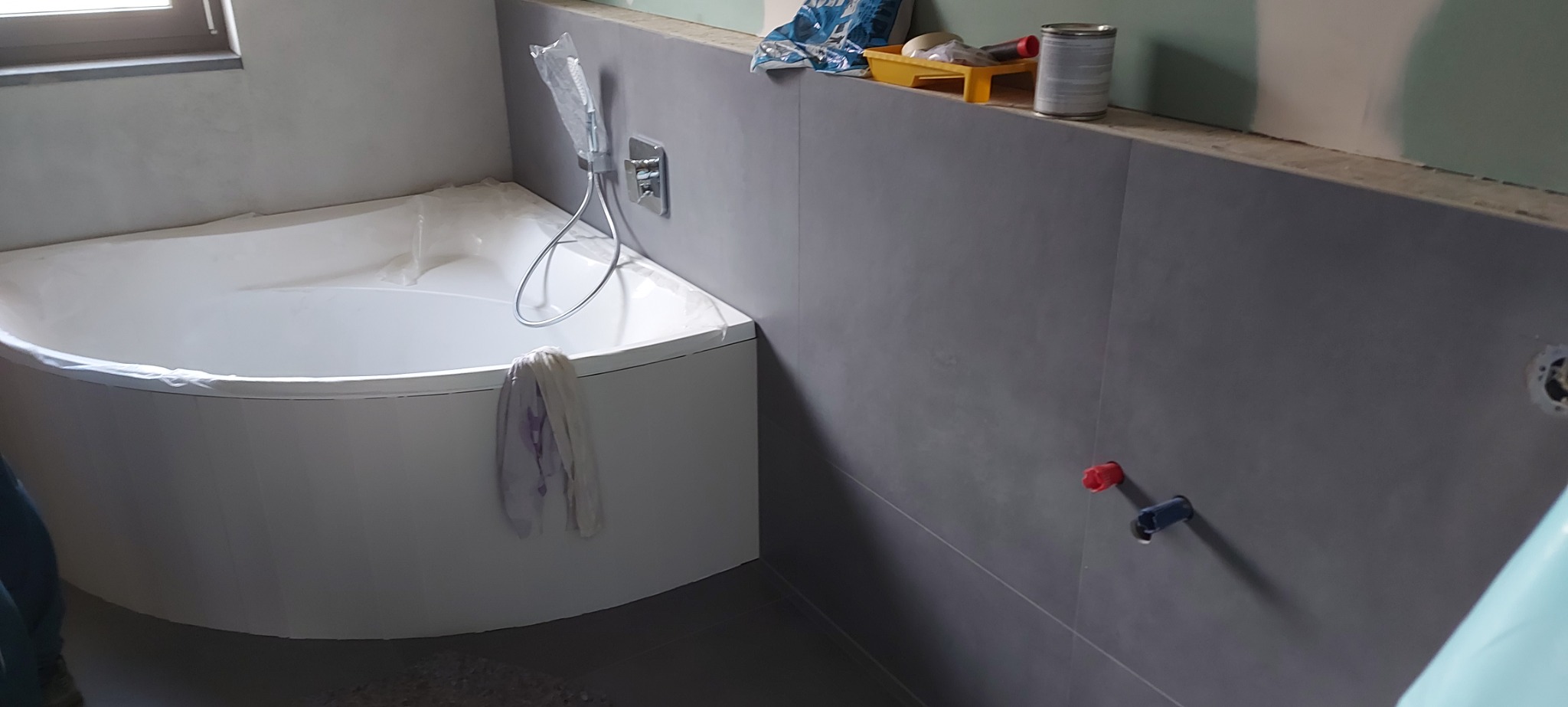 Biała narożna wanna zabezpieczona folią podczas prac wykończeniowych w łazience, widoczne szare płytki ścienne, akcesoria malarskie na górnej półce i wystające rury instalacji wodnej.