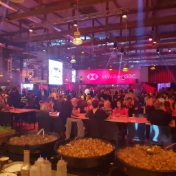 Uroczysta kolacja firmowa z bufetem cateringowym: duże patelnie z jedzeniem na pierwszym planie, goście siedzący przy stołach w tle, oświetlenie eventowe.