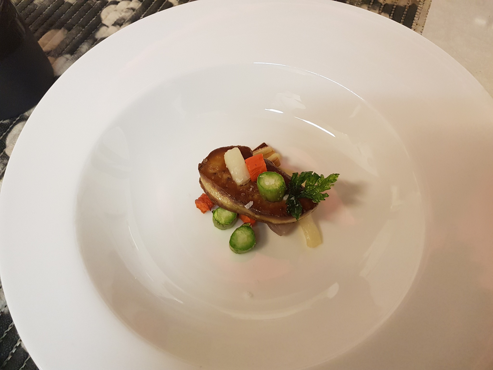Elegancko podane foie gras na białym, głębokim talerzu, udekorowane brukselką, pietruszką i warzywami w jasnych kolorach.