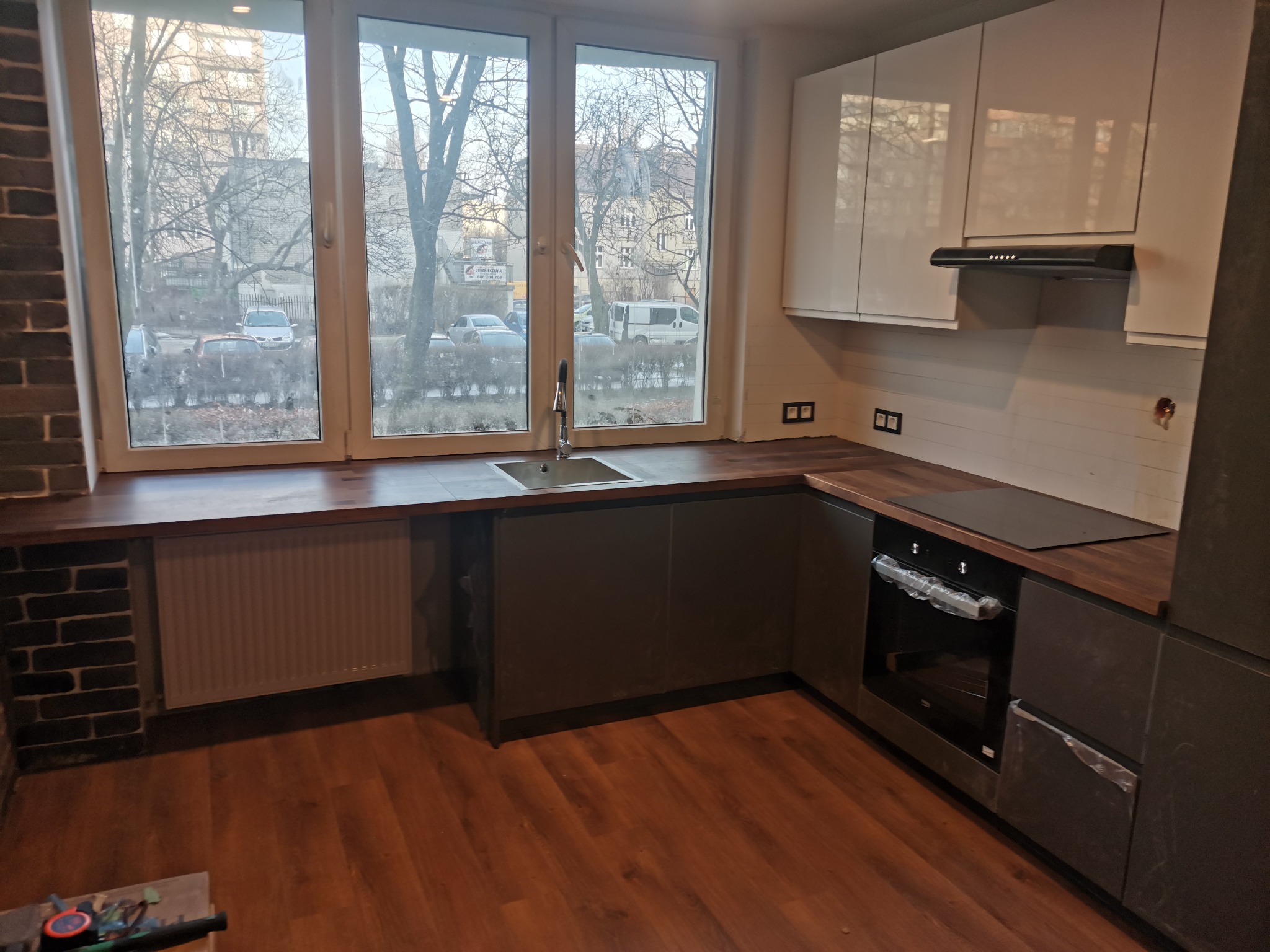 Nowoczesna kuchnia z szarymi szafkami dolnymi i białymi górnymi, drewniany blat, okno z widokiem na drzewa bez liści, piekarnik z folią ochronną na drzwiach, ceglana ściana po lewej stronie.