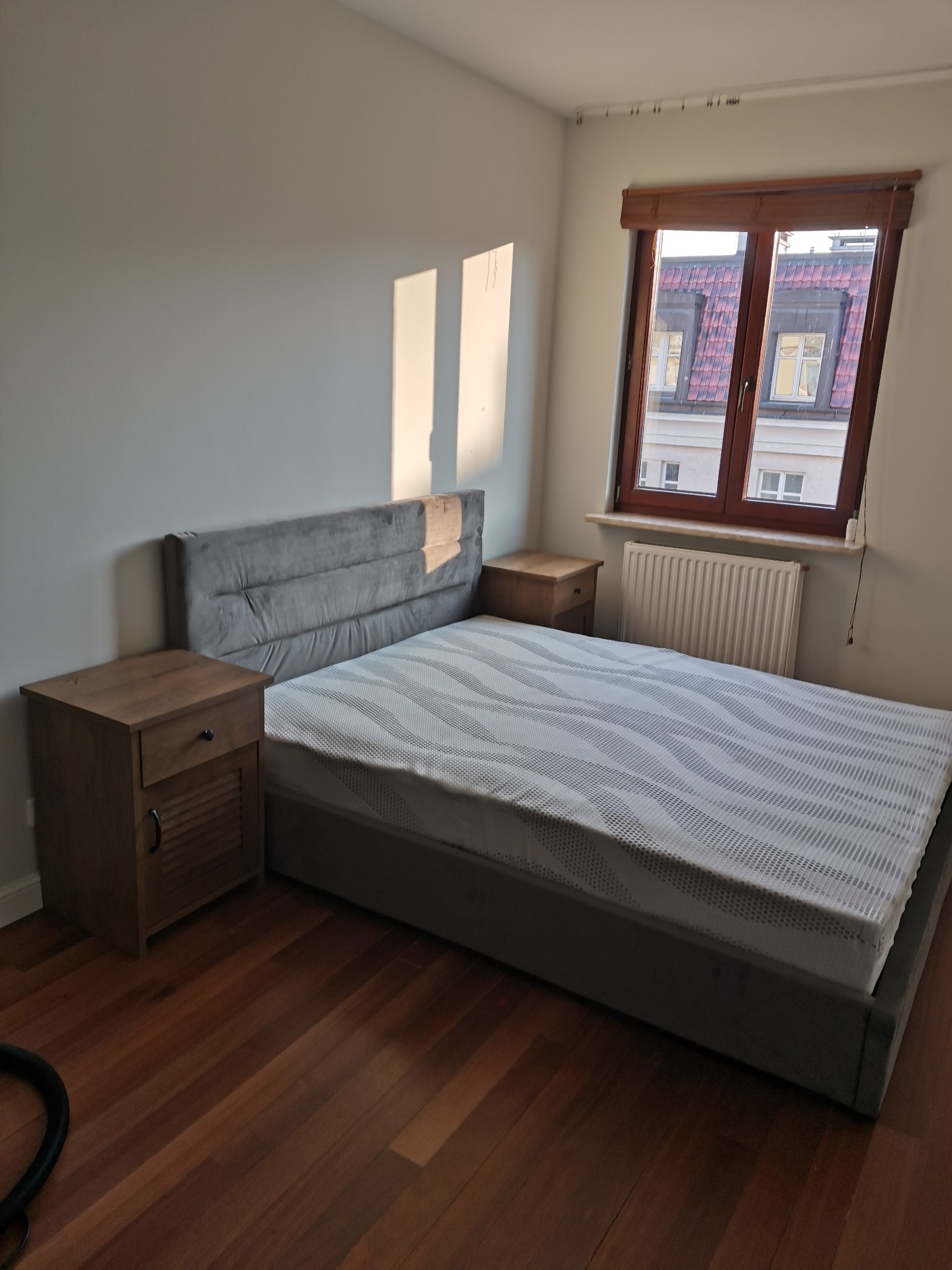 Sypialnia z łóżkiem z szarym wezgłowiem, drewnianymi szafkami nocnymi i parkietem, oświetlona popołudniowym słońcem wpadającym przez okno.