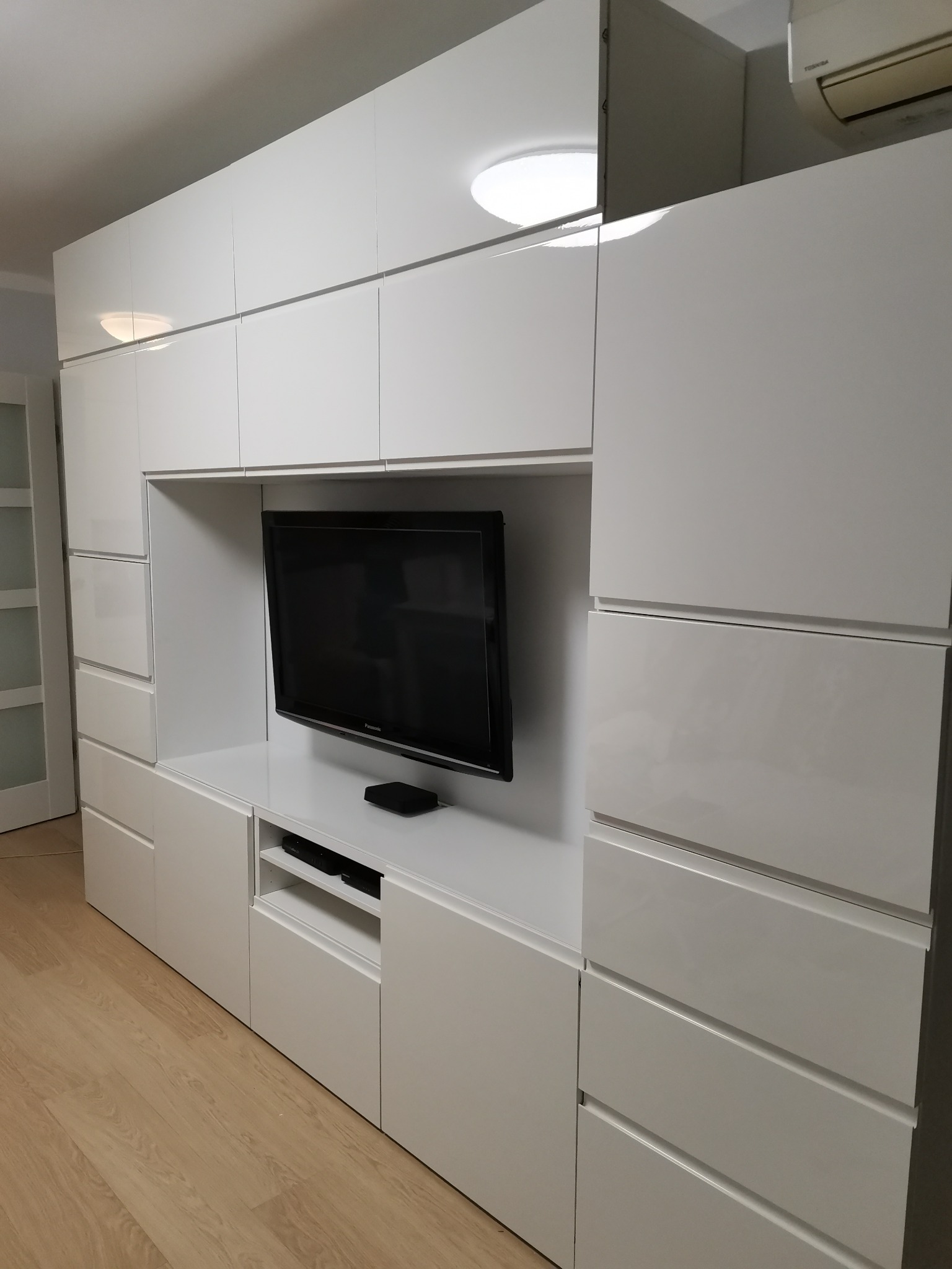 Biała, lakierowana zabudowa meblowa z miejscem na telewizor i licznymi szufladami, oświetlona lampą sufitową, na podłodze panele.