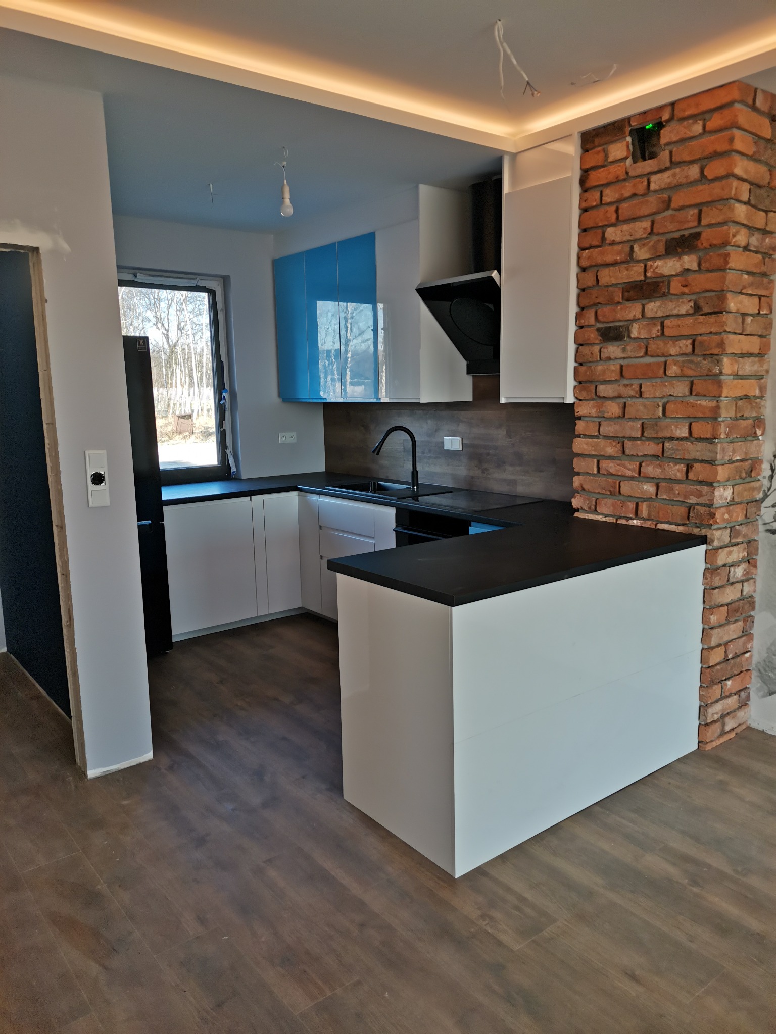 Nowoczesna kuchnia z białymi szafkami i czarnym blatem, akcentem błękitnych szafek wiszących i ceglanym filarem, widoczne niedokończone wykończenia sufitu.