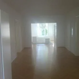 Puste wnętrze po remoncie: parkiet w jodełkę, białe ściany i szafy wnękowe, widok na jasne pomieszczenie z oknem.