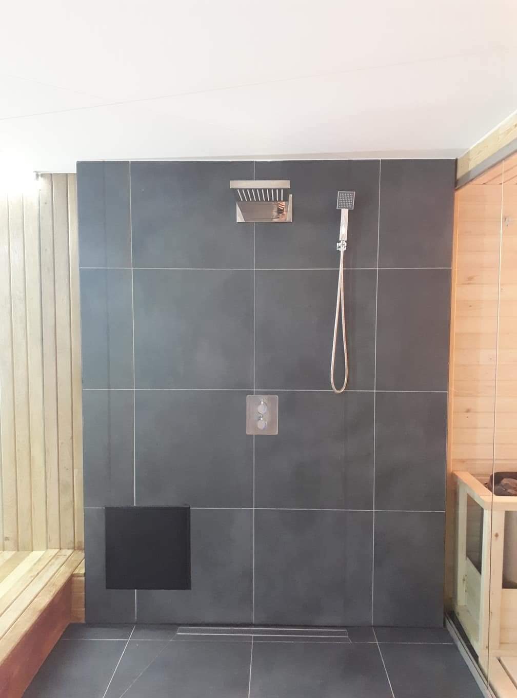 Szara, kwadratowa kabina prysznicowa z deszczownicą i słuchawką, wykończona drewnianą boazerią i ławką w saunie, z widocznym odpływem liniowym.