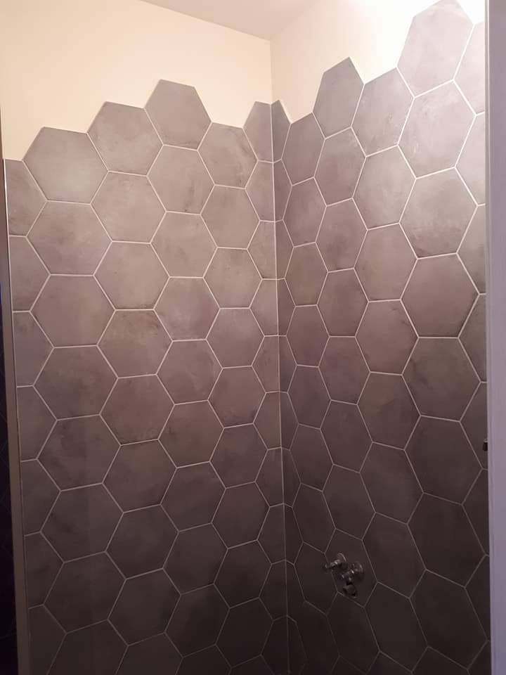 Wykończona ściana prysznica z szarymi, heksagonalnymi płytkami, z widoczną baterią prysznicową i fragmentem białej ściany ponad kafelkami.